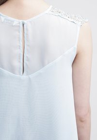 Valkoinen sininen hihaton paita, jossa on läpikuultava selkä, pitsikoristeet olkapäillä ja avaus kaulan takana. Sileä koostumus.