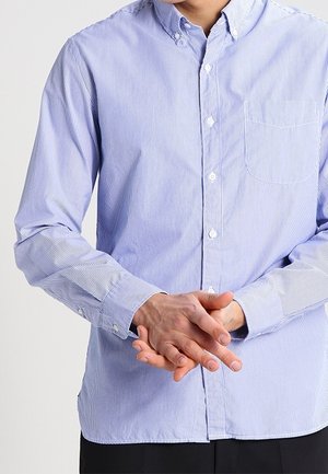 Personne portant une chemise rayée bleu clair à boutons, manches baissées, mains jointes devant à hauteur de la taille.