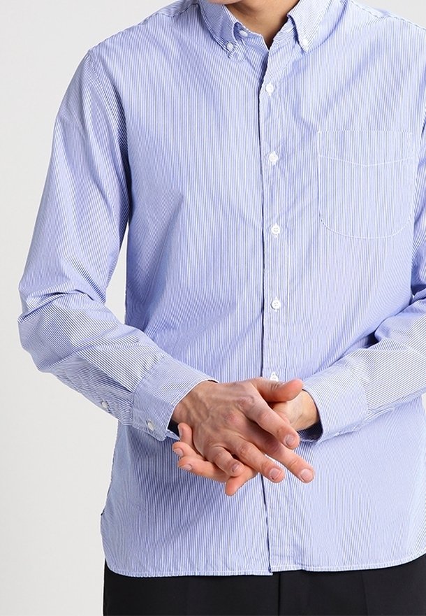 Personne portant une chemise rayée bleu clair à boutons, manches baissées, mains jointes devant à hauteur de la taille.