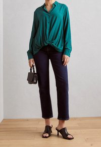 Camicia a maniche lunghe color turchese con bottoni e nodo frontale, abbinata a jeans blu scuro e mule con tacco aperto neri. Porta una piccola borsa nera.