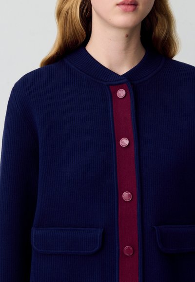 Cardigan a coste navy con listini bordeaux, colletto rotondo e due tasche anteriori. I bottoni sono tondi e di colore rosa.