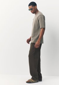 Brown patterned short-sleeve shirt, olive green wide-leg pants, and dark sandals. Model wears dark sunglasses, facing left.