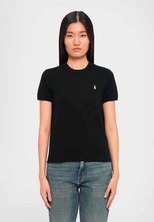 Femme avec des cheveux raides et foncés portant un t-shirt noir à manches courtes avec un petit logo blanc brodé et un jean bleu, se tenant devant un fond blanc.