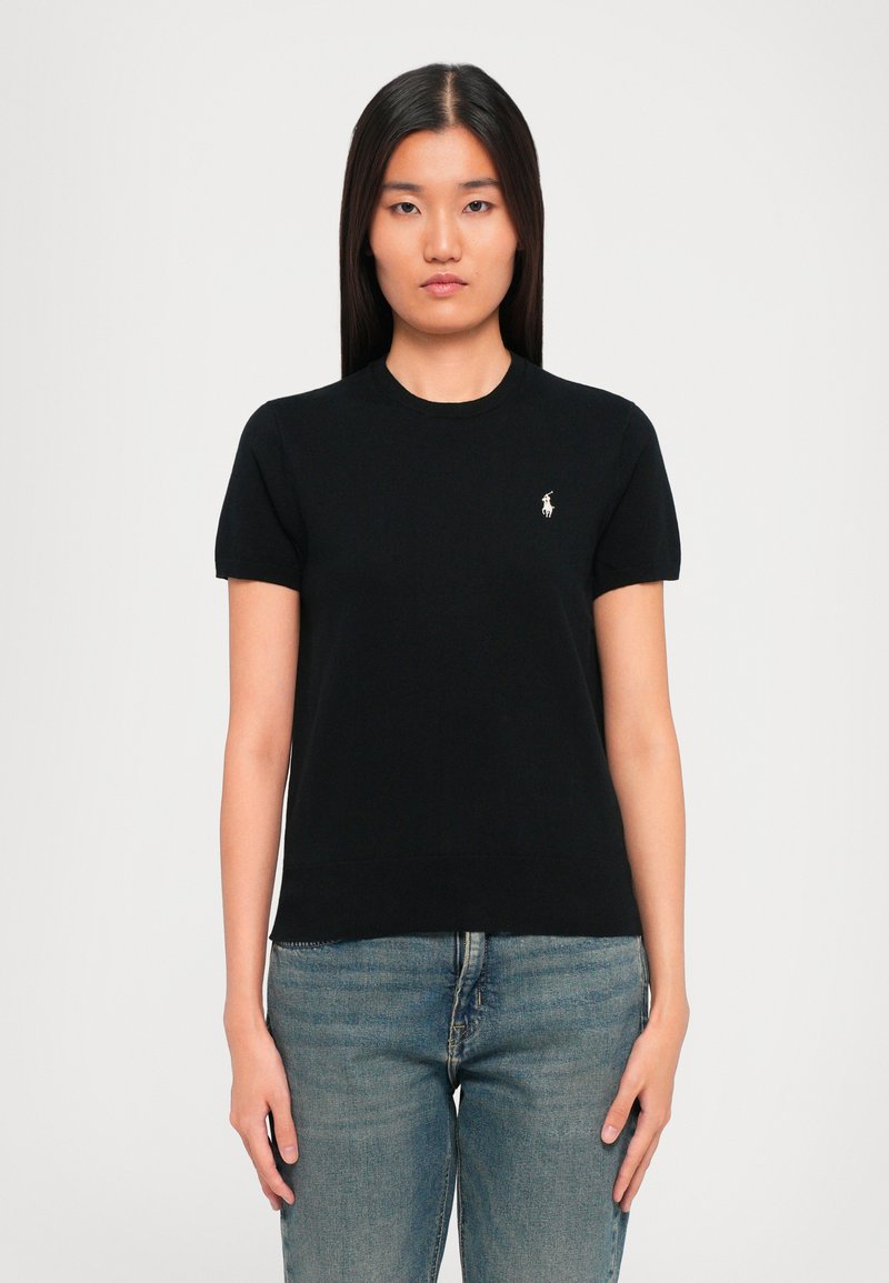 Woman with straight dark hair wearing a black short-sleeve shirt with a small white embroidered logo and blue jeans, standing against white background.