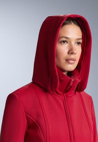 Piros cipzáras dzseki nagy kapucnival, texturált anyagból és puha polár béléssel. A testén átlós steppelt minták találhatók.