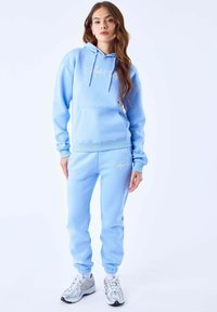 Sudadera de algodón de color azul claro con bolsillo tipo canguro, con cordones y logotipo en el frente. Combinada con pantalones de chándal a juego, también de color azul claro.