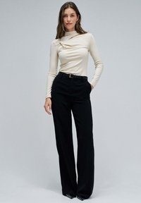 Top beige de manga larga con detalle fruncido en el escote, combinado con pantalones negros de pierna ancha y tacones negros. Textura suave y silueta ajustada.
