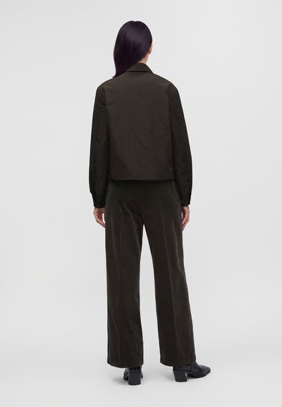 Personne aux longs cheveux foncés portant une veste vert foncé, un pantalon large assorti et des chaussures noires à talons, debout de dos sur un fond uni.