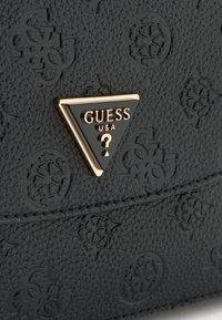 Superficie in pelle nera testurizzata con motivi floreali in rilievo e un distintivo triangolare nero di Guess USA con bordo dorato.