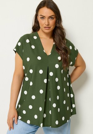 Kvinde med langt brunt hår iført grøn kortærmet bluse med store hvide polkaprikker og blå jeans, stående med den ene hånd på hoften.