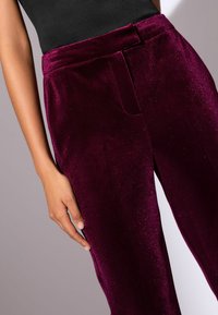 Burgundi velúr nadrág sima textúrával, derékrésszel és oldalsó zsebekkel. Az anyag diszkrét csillogással rendelkezik.