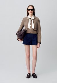 Chaqueta corta de pata de gallo marrón con pajarita blanca, pantalones cortos de mezclilla y mocasines oscuros. La modelo sostiene un bolso de mano marrón oscuro. Lleva gafas de sol cuadradas.