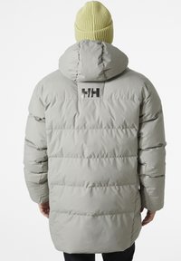 Grijze pufferjas met horizontale stiksels, capuchon en zwart logo detail op de achterkant. Een geribbelde gele beanie wordt gedragen.