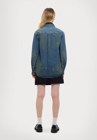 Cămașă din denim albastru cu mâneci lungi și tiv curbat; prezintă un gradient estompat, stropi de vopsea albă și un design clasic al gulerului.