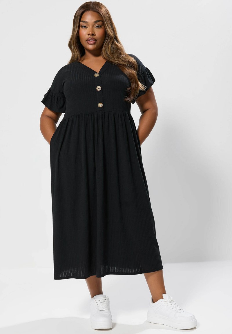 Czarna midi sukienka o prążkowanej teksturze, z krótkimi falbaniastymi rękawami, dekoltem w serek i trzema guzikami z przodu. Noszona z białymi trampkami.