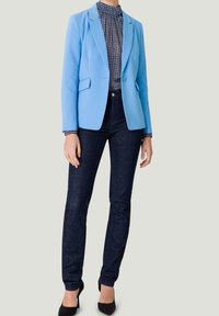 Blazer bleu avec une coupe ajustée, associé à un col roulé à motifs et un jean en denim foncé. Des escarpins noirs complètent l'ensemble.