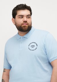 Šviesiai mėlynas polo marškinėlis, pagamintas iš minkštos medvilnės, turintis klasikinius apykaklę, mygtukų užsegimą ir spausdintą apvalų logotipą ant krūtinės.