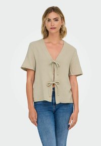 Kortærmet, v-hals top i lys beige tekstureret stof, med en frontknude lukning og afslappet pasform. Båret med blå jeans.