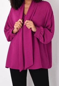 Blouse fuchsia à manches longues avec un col froncé et un détail de nœud à l'avant, associée à un pantalon noir. Tissu lisse, coupe ample et poignets.