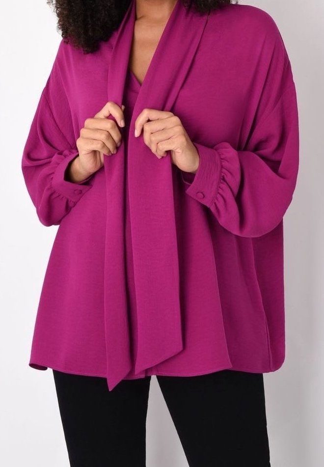 Blouse fuchsia à manches longues avec un col froncé et un détail de nœud à l'avant, associée à un pantalon noir. Tissu lisse, coupe ample et poignets.