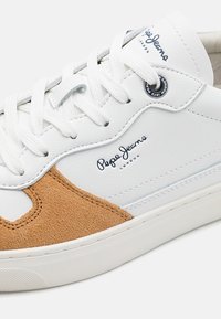 Bílé tenisky s akcenty z semišové kůže v odstínu tan, plochými tkaničkami a gumovou podrážkou. Branding viditelný na boční straně a na jazyku v modré barvě.