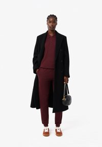 Lång svart kappa över en vinröd polo och matchande byxor, ihop med bruna sneakers och en liten svart handväska i rund form.