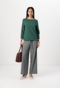 Grön långärmad topp med rund hals, kombinerad med grå vida byxor. En brun handväska och krämfärgade klackskor fulländar outfiten.