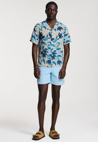 Kortärmad skjorta med knappar i ett mönster av ljusblå, krämfärgade och marinblå blad, kombinerad med ljusblå shorts och bruna slidesandaler.