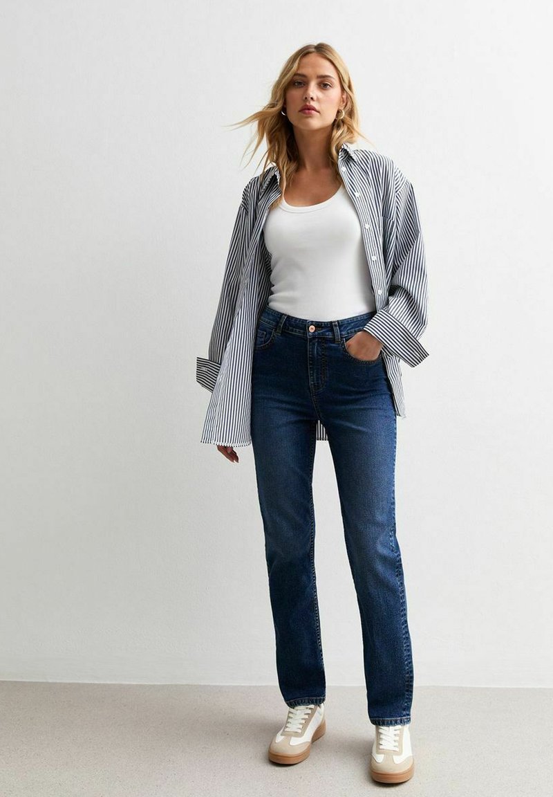 Camisa de botones a rayas en blanco y azul, usada sobre una camiseta sin mangas blanca, combinada con jeans oscuros y zapatillas beiges.