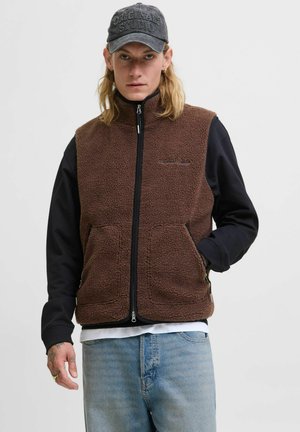 Man wearing a brown fleece zip-up vest over a black long-sleeve shirt, blue jeans, and a dark cap with "Originals Studio" text.