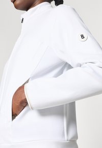 Hvit zip-up jakke med høy krage, teksturerte mesh sider, og en rund logo-patch på ermet. Har innfelt lommer og kontrasterende kant.