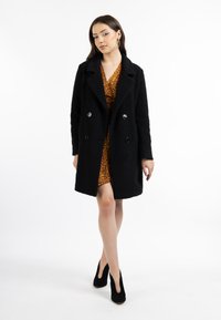 Sort dobbeltradet frakke med tekstureret stof og sølvknapper, lagdelt over en knælængde orange leopardprint kjole og sorte højhælede sko.