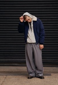 Navy bomberska jakna iz korduroja s kremno ovratnikom, preko črtaste kapucarice. Sive široke hlače z belimi stranskimi črtami in črnimi čevlji.