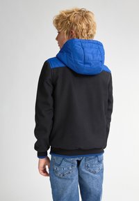Giacca nera con cappuccio blu e dettagli sulle spalle, polsini elasticizzati e una texture liscia. Abbinata a jeans di denim blu.