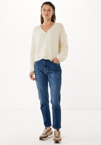 Beige stickad kofta med V-hals och knappar, kombinerad med blå jeans och bruna sneakers med överdel i mocka och mesh.