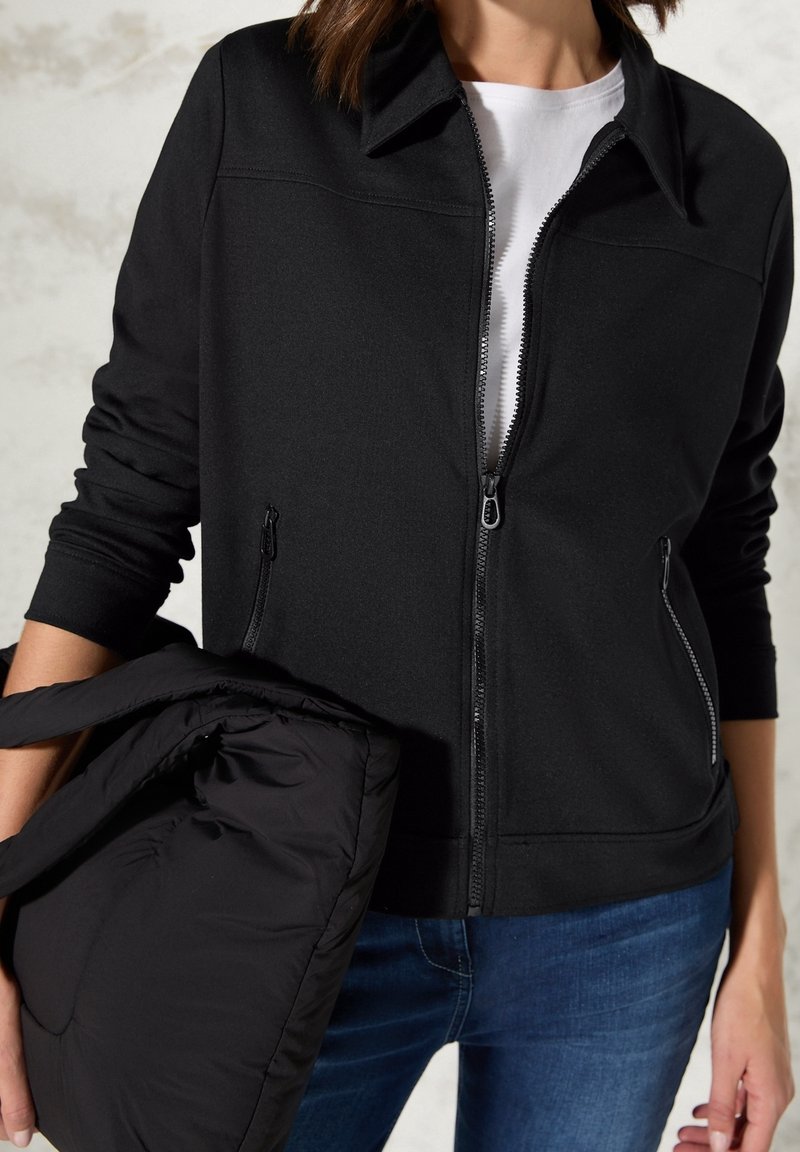 Person in einer schwarz-zipper Jacke über einem weißen Hemd und blauen Jeans, hält eine gefaltete schwarze Daunenjacke in einer Hand.