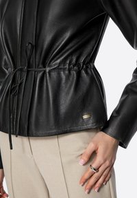 Giacca di pelle nera con dettagli in vita arricciati, maniche lunghe e un piccolo tag del marchio. I pantaloni sono in tessuto chiaro. Mano decorata con un anello.