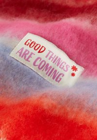 Farverigt, struktureret stof med farvegradienter i pink, rød og lavendel; har et hvidt mærke med teksten "GODE TING KOMMER" i pink og rød.