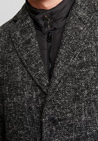 Manteau texturé gris avec une doublure intérieure noire et un col haut. Comprend deux boutons au niveau du col et deux boutons sur le devant.