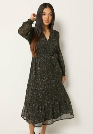 Frau mit langen, glatten Haaren, die ein schwarzes, durchsichtiges Langarmkleid mit braunen Flecken und einem Taillengürtel trägt, vor einem schlichten weißen Hintergrund stehend.