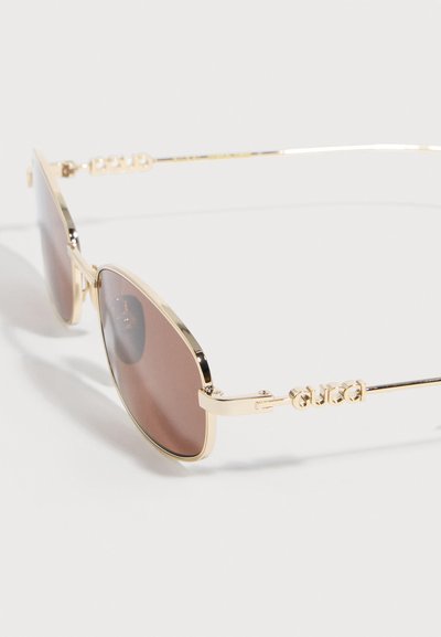 Occhiali da sole con montatura in metallo dorato, lenti sfumate marroni e logo "Gucci" sulle sottili stanghette, mostrati su uno sfondo bianco.