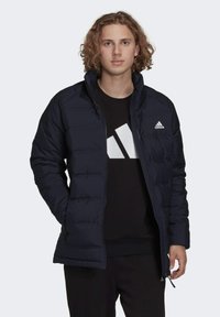 Giubbotto imbottito blu marina con zip frontale, cuciture definite e un piccolo logo bianco sul lato sinistro del petto, indossato sopra una maglietta nera.