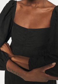 Musta mekko, jossa on neliönmallinen pääntie, kerätty vartalo ja puffihihat. Silkkinen kangas, jossa on hienovarainen tekstuuri, luoden rennon istuvuuden.