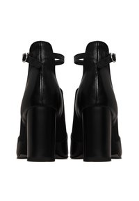 Escarpins noirs en cuir avec bride autour de la cheville et talons carrés épais, vus de dos sur un fond blanc.