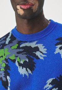 Gros plan sur une personne atteinte de vitiligo au bas du visage et au cou, portant un pull en tricot bleu avec un motif floral abstrait en vert, noir et gris.