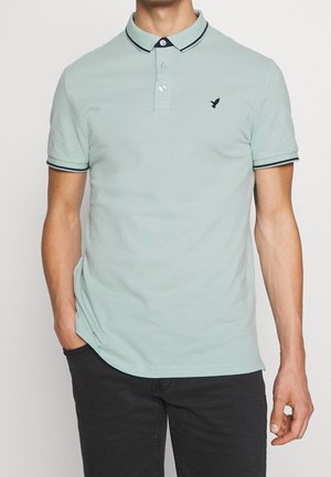 Mann trägt ein hellgrünes Kurzarm-Poloshirt mit schwarzem Besatz an Kragen und Ärmeln, schwarzes Vogel-Logo auf der Brust, Hände in den Taschen einer schwarzen Hose.