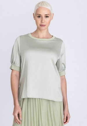 Femme aux cheveux blonds courts portant un chemisier en satin vert clair avec des manches retroussées et une jupe plissée assortie, debout devant un fond uni.