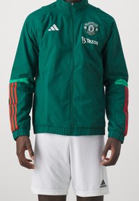 Grøn sportjakke med hvid Adidas-logo, Manchester United-emblem, røde striber på ærmerne og lynlås foran. Hvide shorts med logo.