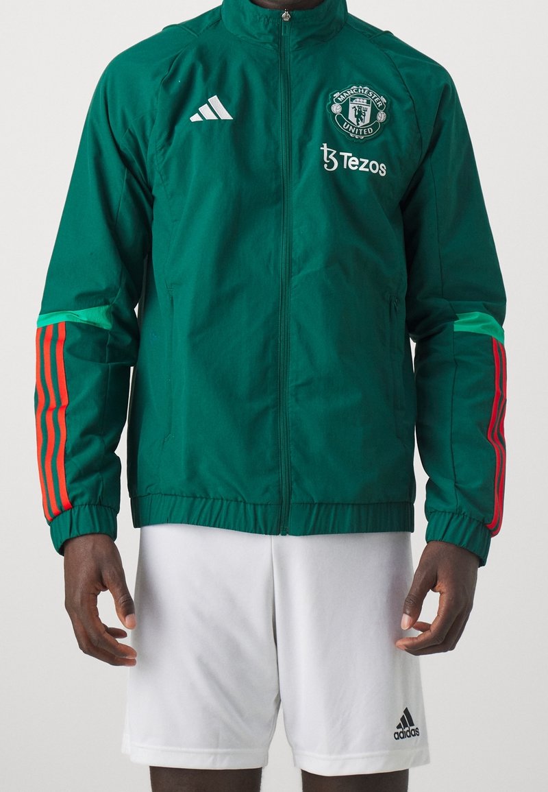 Grøn sportjakke med hvid Adidas-logo, Manchester United-emblem, røde striber på ærmerne og lynlås foran. Hvide shorts med logo.
