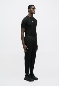 Melnā T-krekls ar logotipa uzliku, melnas bikses ar elastīgu jostasvietu un melnas sporta kurpes. Gluda auduma, pieguļošs dizains un kopumā brīvs piegriezums.